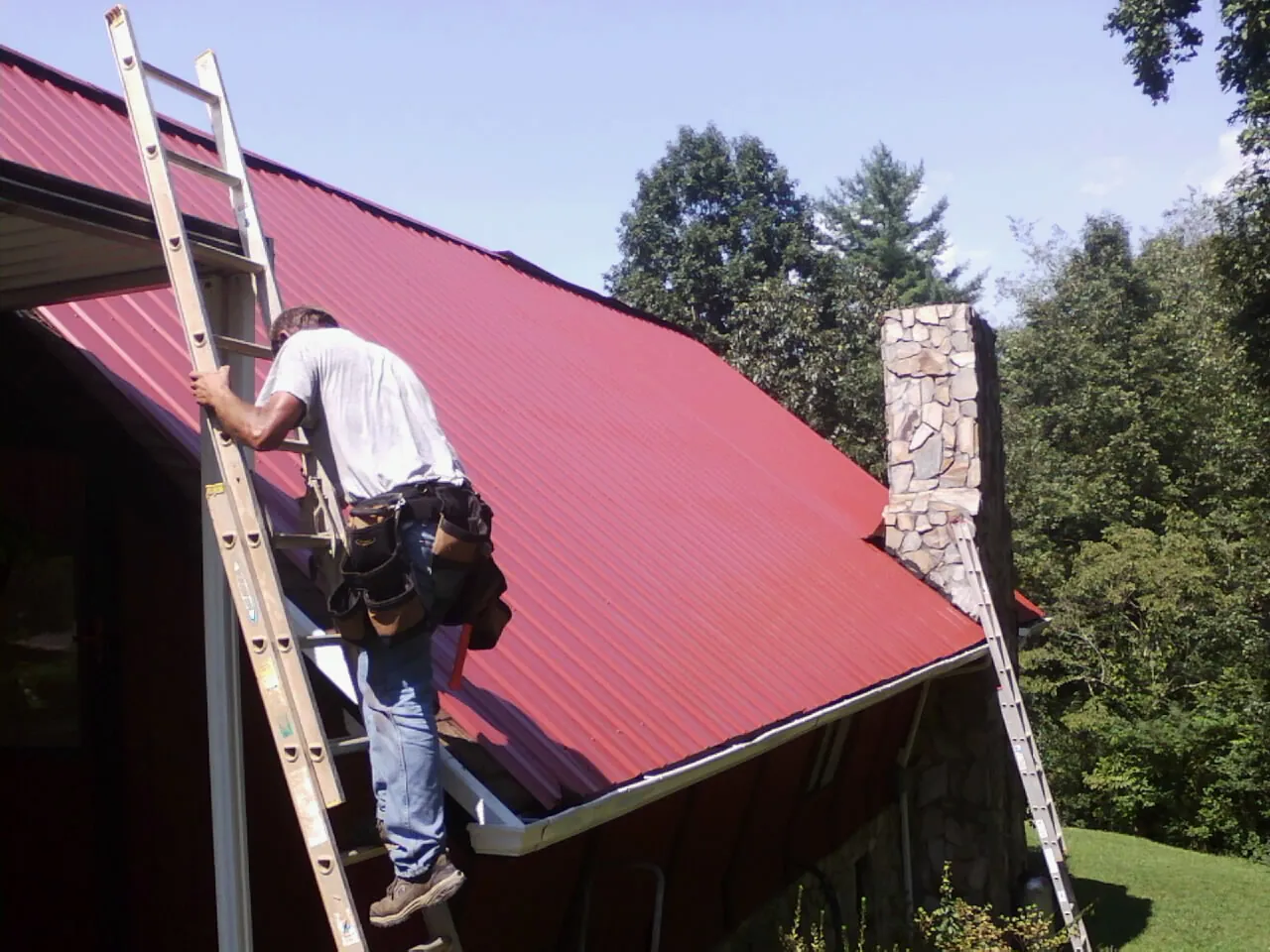 Roofer on a ladder beside a red metal roof and stone chimney — Soffit & Fascia Repair by Roofing Brothers in New Madison
